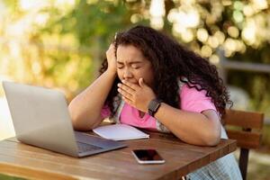 A young woman sits at a wooden table outside, working on her laptop. She appears fatigued and yawning, indicating a long study session in a sunny environment. photo