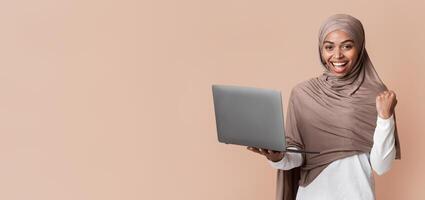 Cheerful Afro Muslim Girl In Headscarf Rejoicing Success With Laptop, Emotionally Raising Fist And Looking At Camera, Yellow Background photo