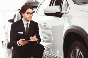 Pensive Auto Insurance Agent Sitting Near New Car Checking It And Taking Notes Working In Automobile Dealership Center photo