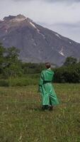 Stylish female model dressed in green leather coat walking in forest on background of volcano on summer day. Vertical . Handheld shot, back view. Part of series video
