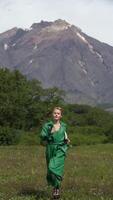 Elegant Caucasian ethnicity female model dressed in green leather coat running in forest against background of volcano on summer weather. Vertical . Handheld shot front view. Part of series video