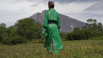 Female model dressed in green long leather coat walking in forest on background of volcano on summer day. Handheld shot, back view. Part of series video