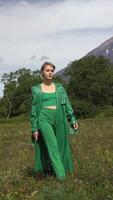 Elegant Caucasian ethnicity female model dressed in green leather coat walking in forest against background of volcano on summer weather. Vertical . Handheld shot front view. Part of series video