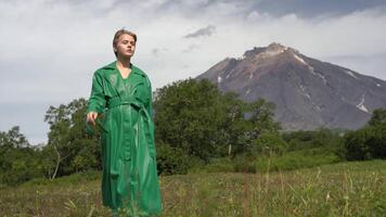 Elegant Caucasian ethnicity female model dressed in green leather coat walking in forest against background of volcano on summer weather. Handheld, front view, low-angle shot. Part of series video