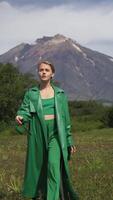 Caucasian ethnicity female model dressed in green top, trousers and leather coat walking in forest on background of mountain on summer day. Vertical . Handheld shot, front view. Part series video