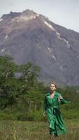 Fashionable woman model dressed in green long leather coat running in forest on background of mountain on summer weather. Vertical , slow motion. Handheld shot front view. Part of series video