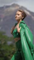 Portrait of fashionable Caucasian female dressed in green top, trousers and leather coat posing in forest on background of volcano on summer day. Vertical , slow motion shot. Part of series video