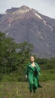Beauty blonde woman model dressed in green top, trousers and leather cloak running in forest on background of mountain on summer weather. Vertical . Slow motion shot front view. Part series video