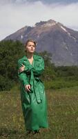Elegant Caucasian ethnicity female model dressed in green leather coat walking in forest against background of volcano on summer weather. Vertical . Handheld shot front view. Part of series video