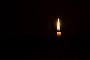 A single burning candle flame or light glowing on a small orange candle on black or dark background on table in church for Christmas, funeral or memorial service with copy space photo