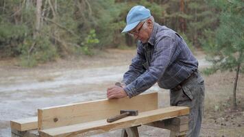 An experienced craftsman carefully processes a wooden board using an old plane. Surrounded by tall trees and natural light, he focuses on his work and enjoys the manual labor. video
