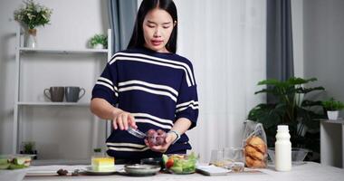 Handheld shot, Young Asian woman prepare food and fruit into cup and bowl on table ready for lunch at home. Lifestyle at home video