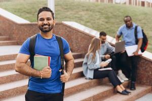 Confident Indian Student Standing with Books on Campus Steps with Group Study Session in Background photo