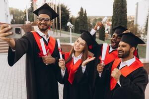 Group of Diverse Graduates Celebrating with Selfie Outdoors in Traditional Caps and Gowns photo