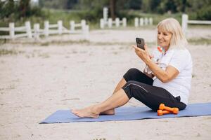 Senior Woman Exercising Outdoors On Beach, Using Smartphone For Online Workout photo