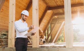 Construction engineer in white helmet. Developer with construction documentation on the background of wooden building under construction. Portrait of smiling architect near eco friendly house. photo