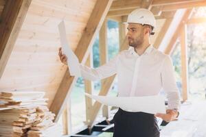 Architect wooden buildings. Man stands under frame wooden house. Man architect studies documentation or blueprints. Guy in protective helmet at construction site. Architect under timber framed house photo