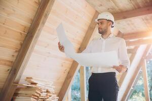 Architect wooden buildings. Man stands under frame wooden house. Man architect studies documentation or blueprints. Guy in protective helmet at construction site. Architect under timber framed house photo