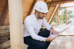 Architect wooden buildings. Man stands under frame wooden house. Man architect studies documentation or blueprints. Guy in protective helmet at construction site. Architect under timber framed house photo