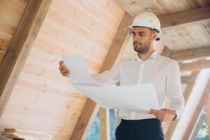 Architect wooden buildings. Man stands under frame wooden house. Man architect studies documentation or blueprints. Guy in protective helmet at construction site. Architect under timber framed house photo