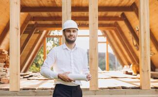 Construction engineer in white helmet. Developer with construction documentation on the background of wooden building under construction. Portrait of smiling architect near eco friendly house. photo