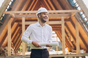 Construction engineer in white helmet. Developer with construction documentation on the background of wooden building under construction. Portrait of smiling architect near eco friendly house. photo