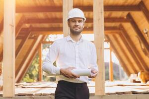 Construction engineer in white helmet. Developer with construction documentation on the background of wooden building under construction. Portrait of smiling architect near eco friendly house. photo