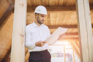 Architect wooden buildings. Man stands under frame wooden house. Man architect studies documentation or blueprints. Guy in protective helmet at construction site. Architect under timber framed house photo