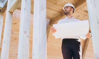 Construction engineer in white helmet. Developer with construction documentation on the background of wooden modular building under construction. Portrait of smiling architect near eco friendly house. photo