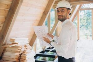 Architect wooden buildings. Man stands under frame wooden house. Man architect studies documentation or blueprints. Guy in protective helmet at construction site. Architect under timber framed house photo