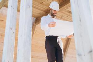 Construction engineer in white helmet. Developer with construction documentation on the background of wooden modular building under construction. Portrait of smiling architect near eco friendly house. photo