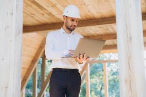 Man builder. Foreman with laptop. Contractor for construction company. Architect in unfinished building. Builder in wooden modular house under construction. Man engineer from construction team photo