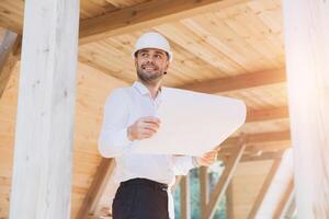 Construction engineer in white helmet. Developer with construction documentation on the background of wooden modular building under construction. Portrait of smiling architect near eco friendly house. photo