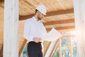 Construction engineer in white helmet. Developer with construction documentation on the background of wooden modular building under construction. Portrait of smiling architect near eco friendly house. photo
