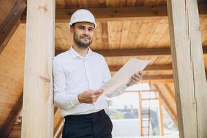 Architect wooden buildings. Man stands under frame wooden house. Man architect studies documentation or blueprints. Guy in protective helmet at construction site. Architect under timber framed house photo