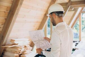 Architect wooden buildings. Man stands under frame wooden house. Man architect studies documentation or blueprints. Guy in protective helmet at construction site. Architect under timber framed house photo