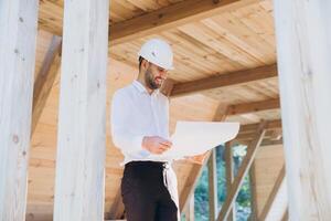 Construction engineer in white helmet. Developer with construction documentation on the background of wooden modular building under construction. Portrait of smiling architect near eco friendly house. photo