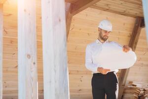 Construction engineer in white helmet. Developer with construction documentation on the background of wooden modular building under construction. Portrait of smiling architect near eco friendly house. photo
