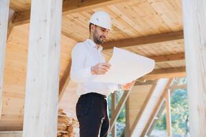 Construction engineer in white helmet. Developer with construction documentation on the background of wooden modular building under construction. Portrait of smiling architect near eco friendly house. photo