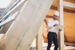 Architect wooden buildings. Man stands under frame wooden house. Man architect studies documentation or blueprints. Guy in protective helmet at construction site. Architect under timber framed house photo