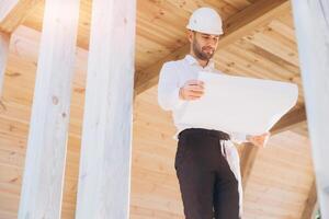Construction engineer in white helmet. Developer with construction documentation on the background of wooden modular building under construction. Portrait of smiling architect near eco friendly house. photo