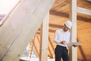 Architect wooden buildings. Man stands under frame wooden house. Man architect studies documentation or blueprints. Guy in protective helmet at construction site. Architect under timber framed house photo