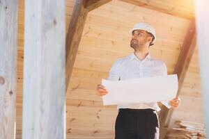 Construction engineer in white helmet. Developer with construction documentation on the background of wooden modular building under construction. Portrait of smiling architect near eco friendly house. photo