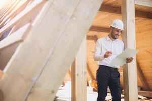 Architect wooden buildings. Man stands under frame wooden house. Man architect studies documentation or blueprints. Guy in protective helmet at construction site. Architect under timber framed house photo