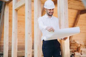 Construction engineer in white helmet. Developer with construction documentation on the background of wooden building under construction. Portrait of smiling architect near eco friendly house. photo