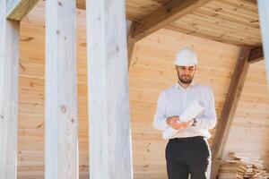 Construction engineer in white helmet. Developer with construction documentation on the background of wooden modular building under construction. Portrait of smiling architect near eco friendly house. photo