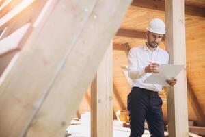 Architect wooden buildings. Man stands under frame wooden house. Man architect studies documentation or blueprints. Guy in protective helmet at construction site. Architect under timber framed house photo
