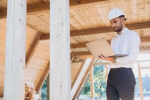 Man builder. Foreman with laptop. Contractor for construction company. Architect in unfinished building. Builder in wooden modular house under construction. Man engineer from construction team photo