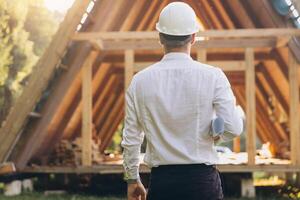Rear view of construction engineer in white helmet. Developer with construction documentation on the background of wooden modular building under construction. photo