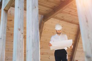 Construction engineer in white helmet. Developer with construction documentation on the background of wooden modular building under construction. Portrait of smiling architect near eco friendly house. photo